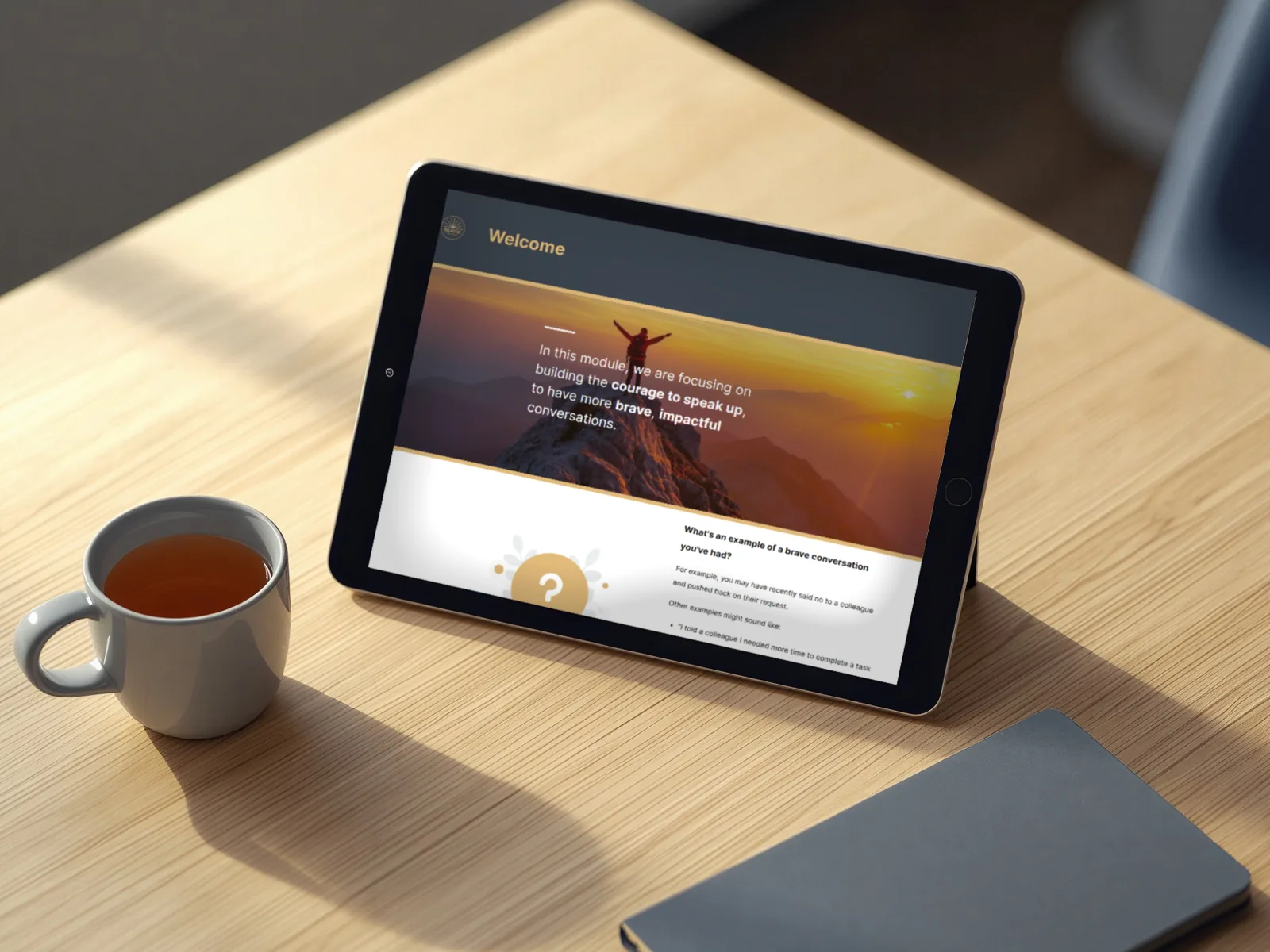 A tablet on a wooden desk showing the welcome page of the Courage to Speak Up module, beside a mug of tea and a notebook
