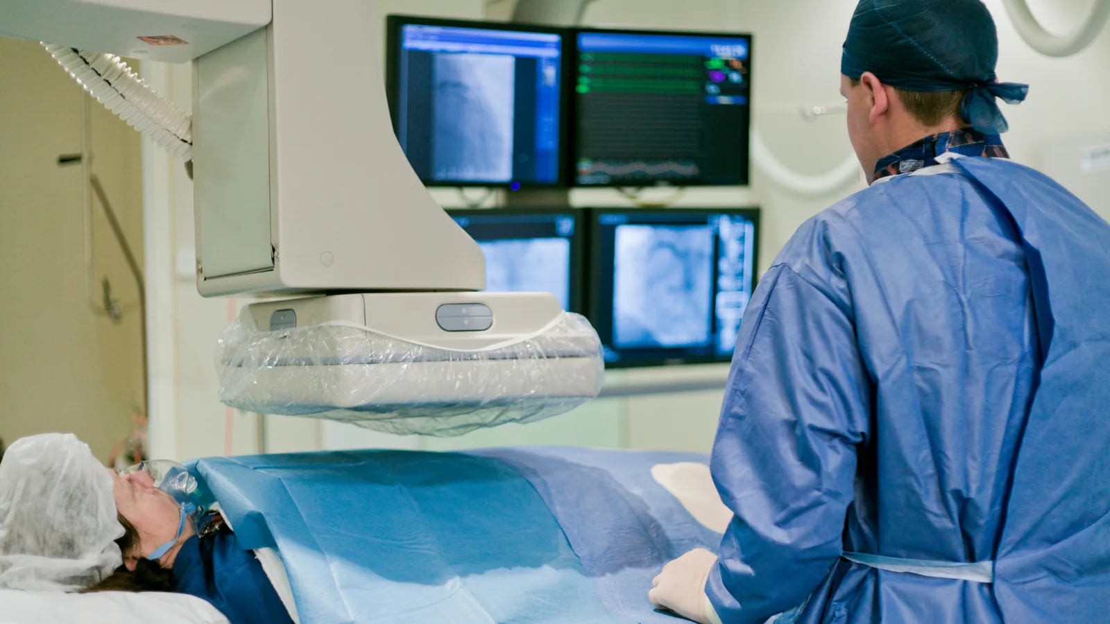 A cardiac catheterisation lab mid-procedure, with imaging screens above the table and a clinician working alongside a draped patient